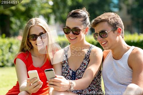 Image of smiling friends with smartphones sitting in park