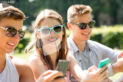 Image of smiling friends with smartphones sitting in park