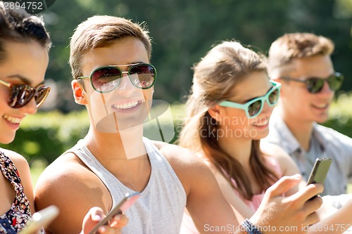 Image of smiling friends with smartphones sitting in park