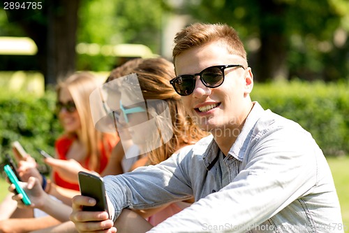 Image of smiling friends with smartphones sitting in park