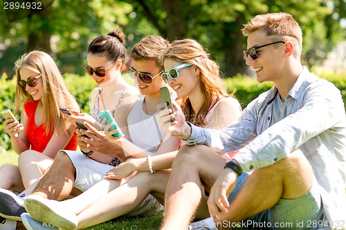 Image of smiling friends with smartphones sitting on grass