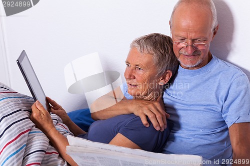 Image of Senior Couple with Tablet and Newspaper in Bed