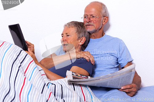 Image of Senior Couple with Tablet and Newspaper in Bed