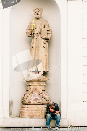 Image of Homeless Old Man Sitting Outdoors Under A Statue Of A Monk In Pr