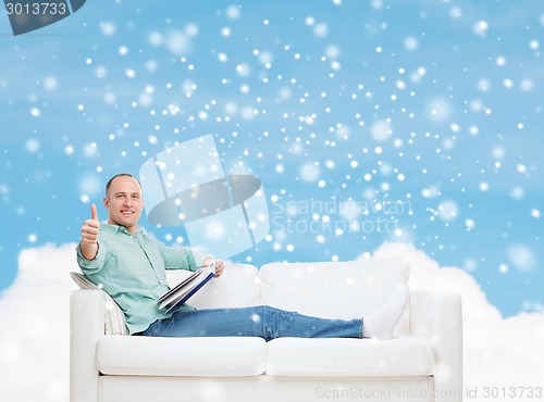 Image of smiling man with book showing thumbs up on sofa
