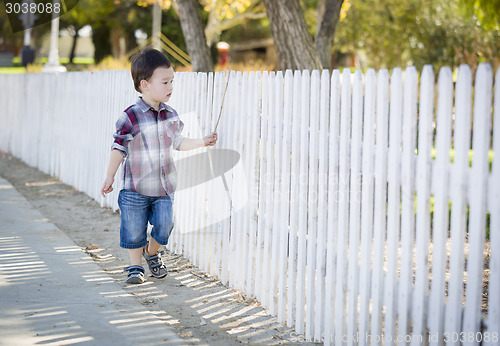 Image of Young Mixed Race Boy Walking with Stick Along White Fence