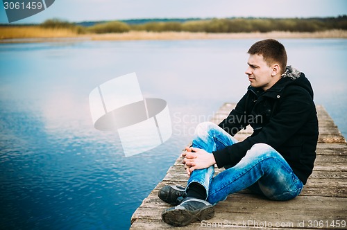 Image of Young handsome man sitting on wooden pier, relaxing,  thinking, 