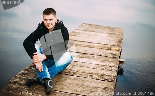 Image of Young handsome man sitting on wooden pier, relaxing,  thinking. 