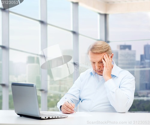 Image of senior man with laptop and pen writing at office