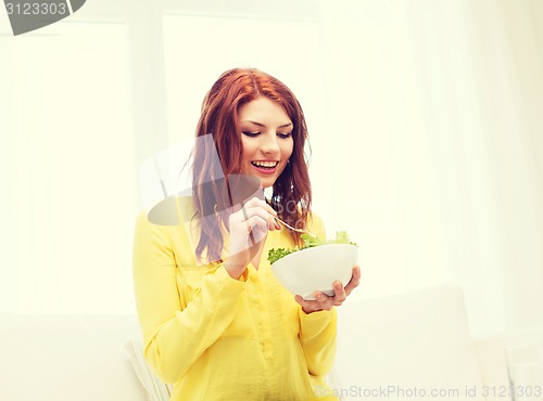 Image of smiling young woman eating green salad at home