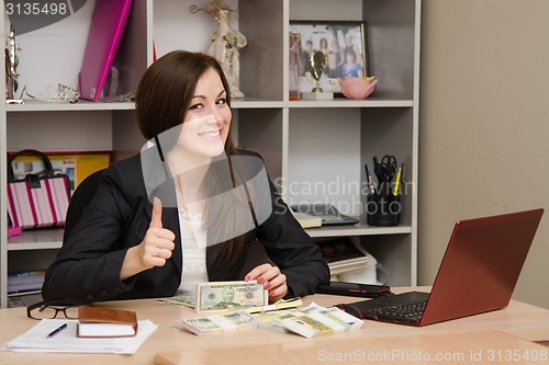 Image of Pretty teenage girl sitting at desk with a pile of money and shows thumb