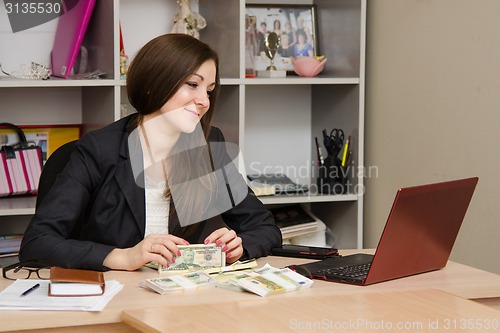 Image of Joyful Girl sitting a desk with pile of money and looking at computer