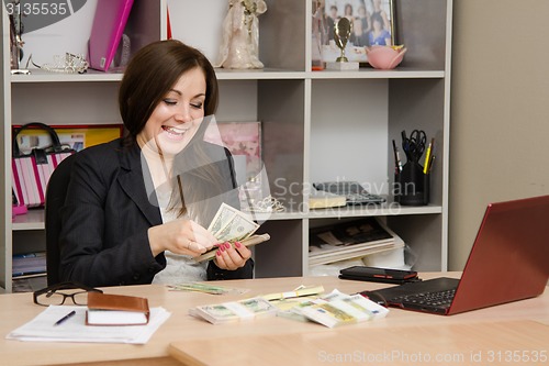 Image of girl behind desk in office feverishly counts banknotes with a triumphant smile