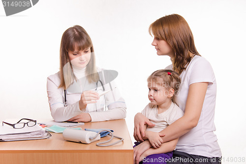 Image of A pediatrician looks at the thermometer, sitting next to a woman with a child on her lap