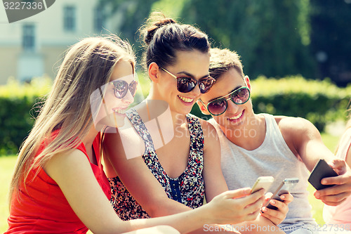 Image of smiling friends with smartphones sitting in park