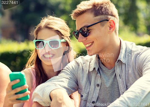 Image of smiling friends with smartphone sitting in park