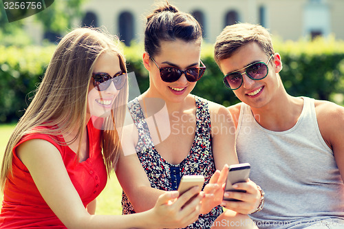 Image of smiling friends with smartphones sitting in park