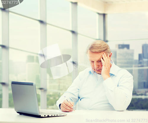 Image of senior man with laptop and pen writing at office