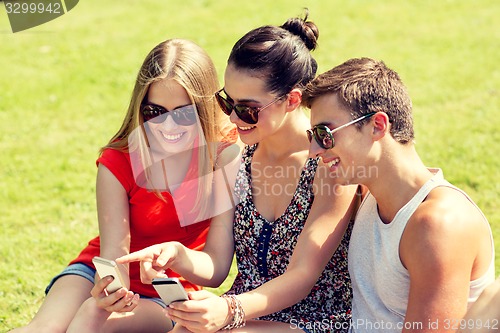 Image of smiling friends with smartphones sitting in park