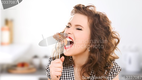 Image of hungry young woman eating meat on fork in kitchen