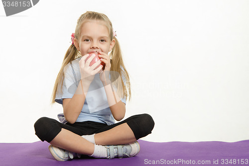 Image of Cheerful girl eating an apple sitting on sport mat