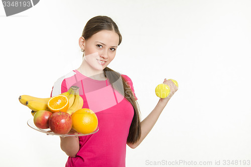 Image of Girl athlete holding a plate of fruit and dumbbell