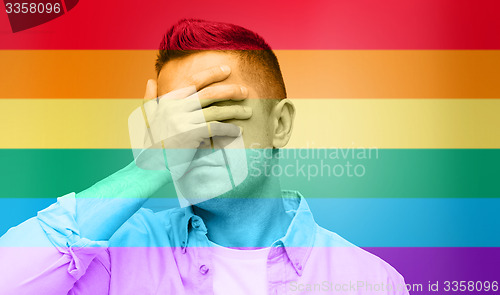 Image of gay covering his face by hand over rainbow flag