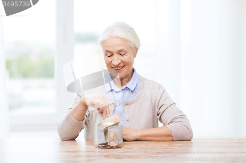 Image of senior woman putting money into glass jar at home