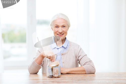 Image of senior woman putting money into glass jar at home