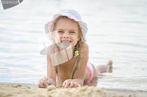 Image of Happy little girl six years lying on the sand on the bank of the river