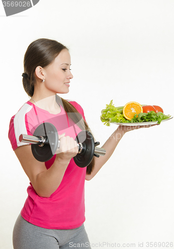 Image of Sportswoman looking at a plate of fruit while holding a dumbbell in the other hand