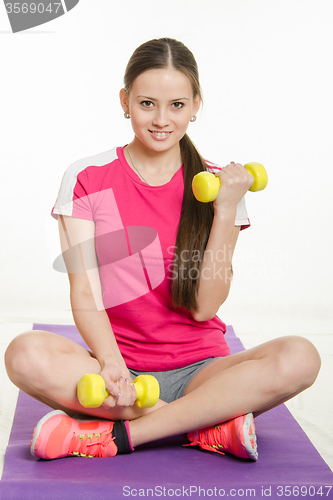Image of Sportswoman sitting on a mat and perform exercises with dumbbells