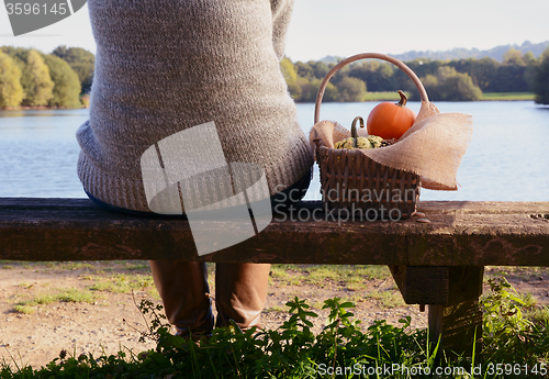 Image of Woman sits on bench with a basket of pumpkins by a lake