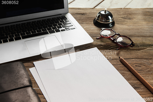 Image of The laptop, blank paper, glasses and small bell on the wooden table 