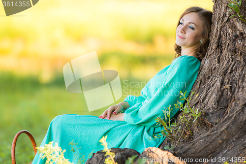 Image of A girl in a long dress and sat resting at a big tree in the forest