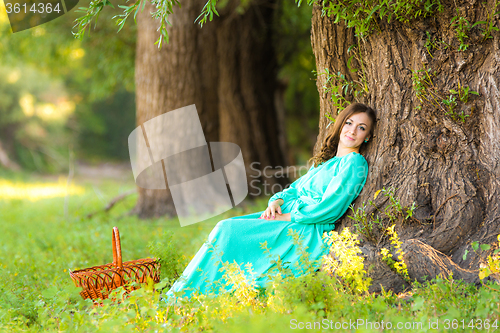 Image of A girl in a long dress and sat resting at a big tree in the forest