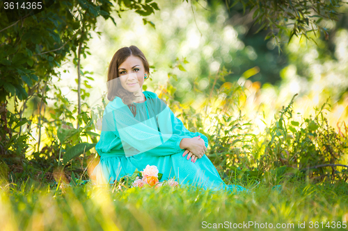 Image of A girl in a long dress sitting on the grass in the forest in sunny weather