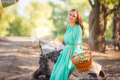 Image of The girl with a basket in a long dress sat down to rest on an old fallen tree