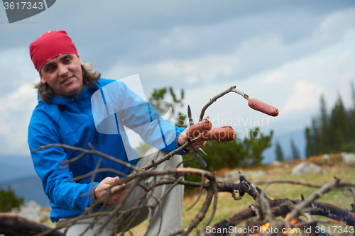 Image of hiking man prepare tasty sausages on campfire