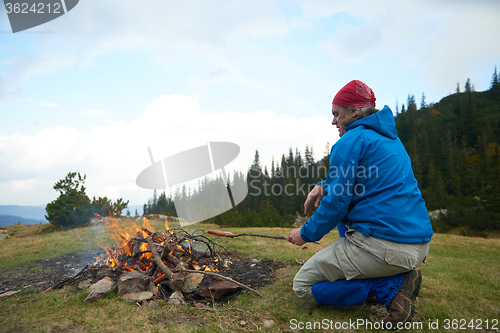 Image of hiking man prepare tasty sausages on campfire