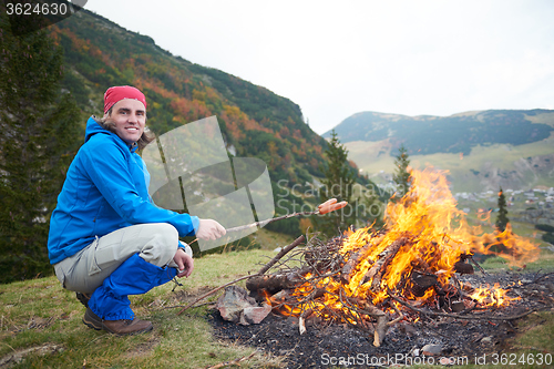 Image of hiking man prepare tasty sausages on campfire