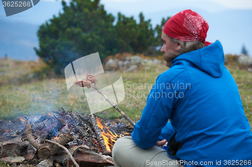 Image of hiking man prepare tasty sausages on campfire