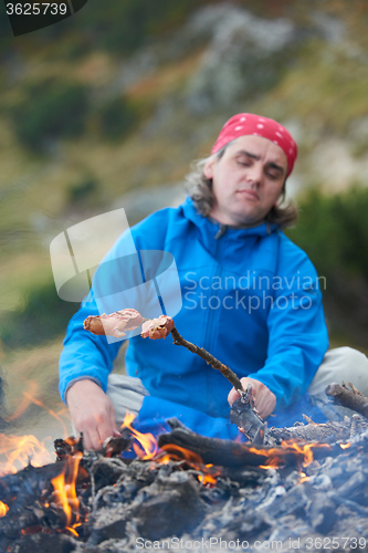 Image of hiking man prepare tasty sausages on campfire