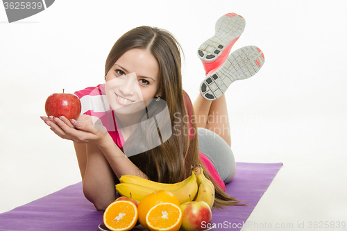 Image of Sportswoman lying on a mat, holding an apple in hand, it is faced with a plate of fruit