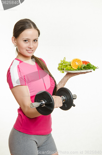 Image of Sportswoman holding a dumbbell and a bowl of fruit