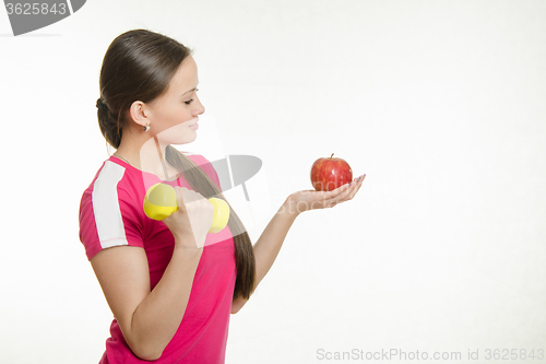 Image of Athlete shakes muscles of the right hand dumbbell and looking at an apple in her left hand