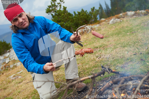 Image of hiking man prepare tasty sausages on campfire