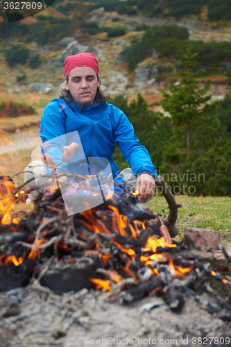 Image of hiking man prepare tasty sausages on campfire