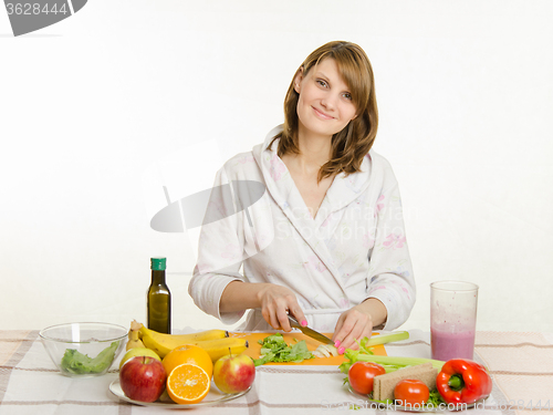 Image of Happy housewife sitting at the table and cut the celery salad