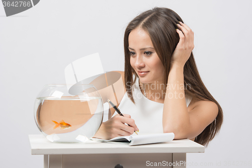 Image of Girl writing in a notebook and looking at a goldfish in an aquarium